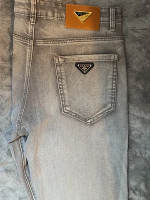 Gråa raka jeans från Prada - Snygga gråa jeans från Prada med raka ben och klassisk femficksdesign. Jeansen har en ikonisk Prada-logga i metall på bakfickan och en läderpatch i midjan. Perfekta för dig som gillar stilrena och exklusiva plagg.