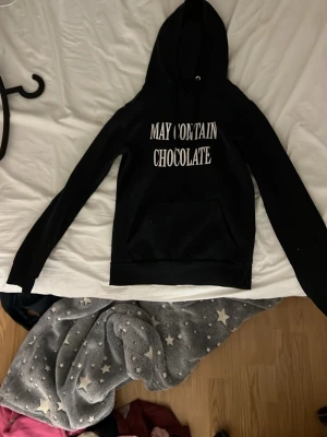Svart hoodie med texttryck - Säljer en svart hoodie med vit text på bröstet där det står 'MAY CONTAIN CHOCOLATE'. Hoodien har huva med dragsko och en klassisk magficka framtill. Tillverkad i mjukt material och har ribbade muddar vid ärmar och nederkant. Perfekt för dig som gillar chill stil.