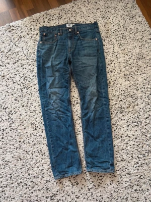 Blå raka jeans från 157 - Säljer ett par klassiska blå jeans från 157 med raka ben och fem fickor. Jeansen har orangea sömmar och silverfärgade nitar. Materialet är slitstark denim i bomull. Perfekta för en avslappnad och tidlös stil.