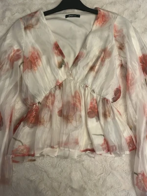 Blommig transparent blus med v-ringning - Säljer en romantisk blus från Gina Tricot i transparent vitt tyg med rosa och röda blommor. Blusen har v-ringning, markerad midja och långa ärmar. Perfekt för dig som gillar en feminin och luftig stil.