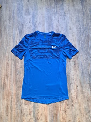 Blå träningstopp från Under Armour - Snygg blå träningstopp från Under Armour med korta ärmar. Överdelen har ett mörkare blått camouflagemönster och logga på bröstet. Tillverkad i ett lätt och snabbtorkande syntetmaterial som passar perfekt till träning.
