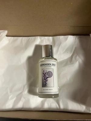 SORENSEN 26.0 Extrait de Parfum - En stilren parfymflaska med metallock och transparent glas. Etiketten har en spiraldesign i lila och svart samt texten 'SORENSEN 26.0' och 'one bold chemist'. Flaskan har en modern och minimalistisk look.