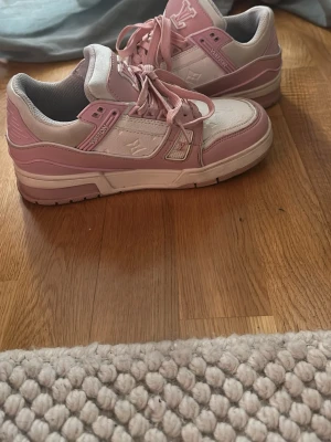 Louis Vuitton rosa och vita sneakers - Säljer ett par snygga Louis Vuitton sneakers i rosa och vitt. Skorna har lågt skaft, klassisk LV-logga på sidan och detaljer i både läder och textil. Sulan är vit med rosa inslag och har tydlig Vuitton-text undertill. Perfekta för dig som vill sticka ut med stil.
