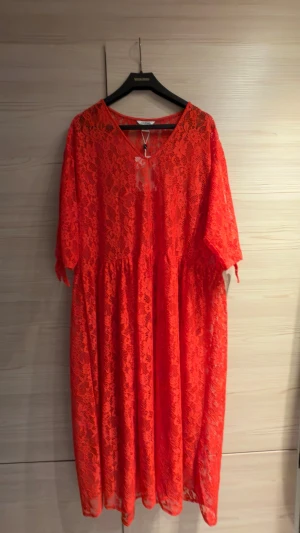 Röd spetsklänning med v-ringning - En röd långklänning i spets med v-ringning och halvtransparent material. Klänningen har knytärmar. Oversized i modellen. Oanvänd med prislapp.
