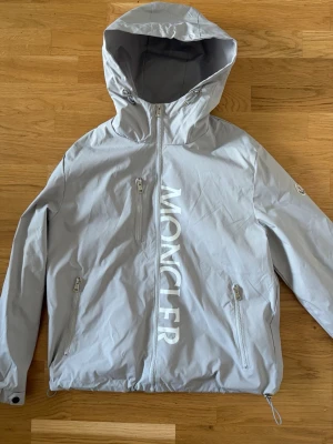 Ljusgrå vindjacka från Moncler - Snygg ljusgrå vindjacka från Moncler med stor logga framtill. Jackan har huva, hel dragkedja, två sidofickor med dragkedja och justerbar nederkant. Tillverkad i lätt och tåligt material, perfekt för blåsiga dagar. Jackan är L men sitter som en M. Inge defekter och aldrig använd.