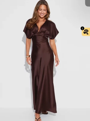 Brun långklänning i satin - Elegant brun långklänning i glansigt satinmaterial med v-ringning och korta, vida ärmar. Klänningen har en smickrande passform och faller snyggt längs kroppen. Perfekt för dig som vill ha en stilren och trendig look.
