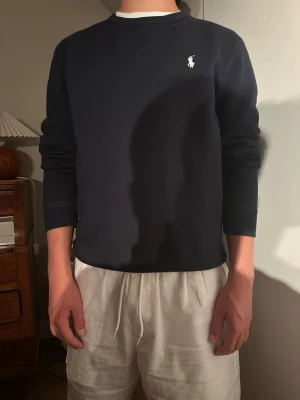 Mörkblå sweatshirt från Polo Ralph Lauren - Klassisk mörkblå sweatshirt från Polo Ralph Lauren med rund halsringning och vit broderad logga på bröstet. Tröjan har långa ärmar och ribbade muddar vid ärmslut och nederkant. Perfekt för en avslappnad och stilren look.