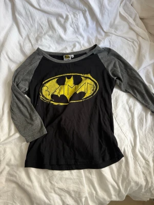 Batman t-shirt med trekvartsärm - Säljer en svart och grå Batman t-shirt med trekvartsärm. Framsidan har en stor gul Batman-logga med sliten look. Perfekt för dig som gillar superhjältar och vill ha en avslappnad stil.
