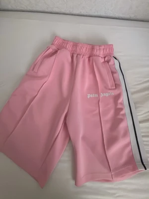 Rosa shorts från Palm Angels - Säljer ett par rosa shorts från Palm Angels med vit logga på ena benet. Shortsen har elastisk midja, sidofickor och en vit rand med svart kant längs sidorna. Perfekta för en sportig och trendig look.