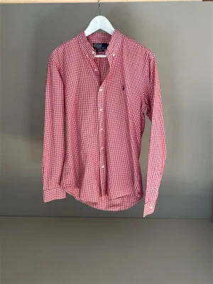 Rödvitrutig skjorta från Polo Ralph Lauren - Klassisk rödvitrutig skjorta från Polo Ralph Lauren med button-down krage och broderad logga på bröstet. Skjortan har långa ärmar, vita knappar och är tillverkad i mjuk bomull. Perfekt för en stilren och avslappnad look.