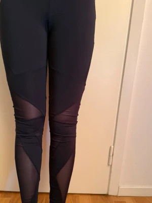 Svarta leggings med meshdetaljer - Snygga svarta leggings med genomskinliga meshpaneler på benen som ger en sportig och trendig look. Tight passform och stretchigt material.