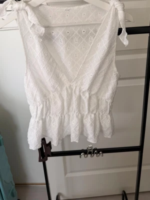 Vit spetslinnetopp med knyt - Söt vit linnetopp i spets med omlott framtill och volang nedtill. Axelbanden knyts i rosetter och tyget har ett diskret broderat mönster. Perfekt för sommarens varma dagar och ger en romantisk vibe.