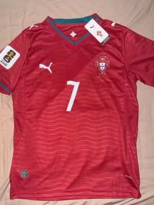 Portugal matchtröja Ronaldo 7 VM 26 - Röd Portugal fotbollströja med nummer 7 och Ronaldo på ryggen. Tröjan har vita detaljer, grönt vid kragen och officiella märken på bröstet och ärmen. Perfekt för dig som älskar fotboll och vill stötta landslaget.