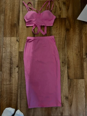 Rosa tvådelat set med crop top - Säljer ett rosa tvådelat set med en vadlång kjol och en crop top med vadderade kupor, smala axelband och dekorativa band framtill. Setet har en tight passform och är gjort i ett stretchigt material som känns mjukt mot huden.