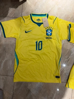 Brasilien Neymar JR matchtröja gul - Gul Brasilien matchtröja från Nike med Neymar JR och nummer 10 på ryggen. Tröjan har blå detaljer, CBF-emblem på bröstet och är tillverkad i lätt, ventilerande material. Perfekt för fotbollsfans som vill stötta landslaget.