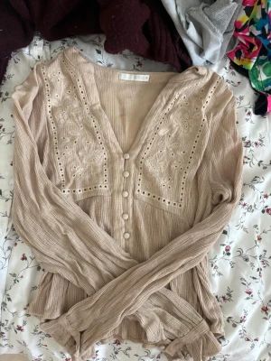 Beige blus med broderi och knappar - Supersöt beige blus med långa ärmar, broderade blommor och dekorativa knappar framtill. Blusen har v-ringning och är gjord i ett mjukt, strukturerat tyg som ger en avslappnad men ändå chic look. Perfekt att styla med jeans eller kjol.