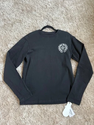 Chrome Hearts tröja - Svart långärmad t-shirt från Chrome Hearts med tryck på bröstet och stor amerikansk flagga med logga och text på ryggen. Skön passform och tillverkad i mjuk bomull. Perfekt för dig som gillar streetwear och vill sticka ut. 