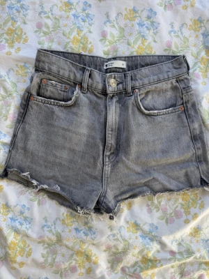 Grå shorts med rå kant - Säljer ett par grå shorts med hög midja och rå, fransig kant nedtill. Klassisk femficksmodell med knapp och dragkedja framtill. 