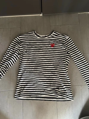 Randig långärmad topp Comme des Garçons - Säljer en svartvit randig långärmad topp från Comme des Garçons Play med det ikoniska röda hjärtat broderat på bröstet. Klassisk rund halsringning och mjukt bomullsmaterial. Perfekt för dig som gillar streetstyle och vill ha något tidlöst.