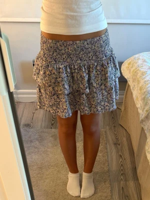 Blommig volangkjol från Gina Tricot -  kort blommig kjol från Gina Tricot i lila, gul och mörkblå. Kjolen har volangdetaljer och smock i midjan för en bekväm passform.  