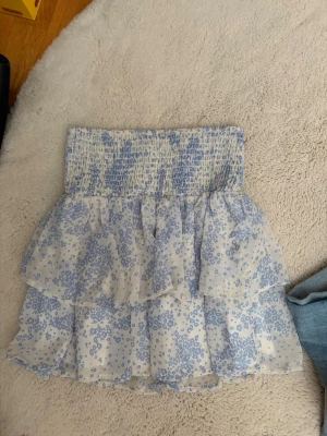 Blommig volangkjol i blått och vitt - Supersöt kort kjol med blått blommönster på vit botten. Kjolen har bred smock i midjan och dubbla volanger som ger en luftig och lekfull känsla. Perfekt för varma dagar och enkel att matcha med en enkel topp.