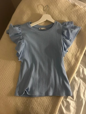 Ljusblå ribbad topp med volangärm - Supersöt ljusblå topp från Zara med ribbad struktur och stora volangärmar. Perfekt för dig som vill ha en feminin och trendig look. Rund halsringning och figurnära passform. Den kan se lite urtvättad ut men inget jag tänker på💓💓