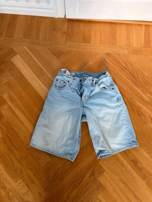 Ljusblå denimshorts från Levis  - Säljer ett par ljusblå denimshorts med klassisk femficksdesign och bälteshällor. Shortsen har en rak passform och knappgylf. Perfekta för varma dagar och ger en avslappnad look.