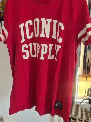 Röd t-shirt med tryck ICONIC SUPPLY - Röd t-shirt med vit text 'ICONIC SUPPLY' på bröstet och vita ränder på ärmarna. Klassisk passform och rund halsringning. Perfekt för en avslappnad och sportig stil.