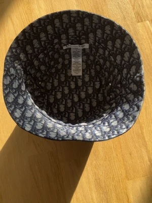 Mönstrad bucket hat från Dior - Säljer en snygg bucket hat från Christian Dior med ikoniskt blått och vitt monogrammönster. Hatten är tillverkad i en mix av polyester, bomull och polyuretan och har en klassisk form med bred kant. Perfekt för dig som vill sticka ut med en exklusiv accessoar.