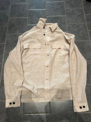 Beige overshirt från Only & Sons - Stilren beige overshirt från Only & Sons med två bröstfickor och svarta knappar. Overshirten har klassisk krage, långa ärmar och knappstängning vid ärmslut. Tillverkad i ett mjukt tyg som passar perfekt för lager på lager. Helt ny aldrig använd.