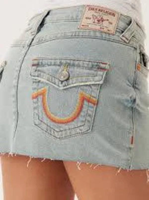 Ljusblå jeanskjol med broderade fickor - Snygg ljusblå jeanskjol med råa kanter och broderade bakfickor i rött och gult. Kjolen har klassiska jeansdetaljer och knappstängning. Perfekt för dig som gillar en avslappnad och trendig stil.