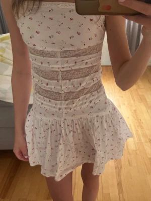 Vit blommig kortklänning med spets - Söt vit kortklänning med små blommor och breda spetsdetaljer över livet. Klänningen har en volangig kjol och är ärmlös med en rak halsringning. Perfekt för sommarens varma dagar och har en romantisk vibe.