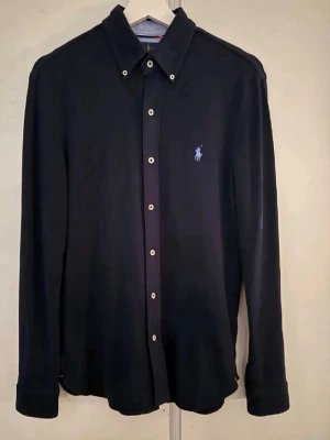 Mörkblå skjorta från Polo Ralph Lauren - Klassisk mörkblå skjorta från Polo Ralph Lauren med knappslå och lång ärm. Skjortan har en broderad logga i ljusblått på bröstet och vita knappar. Perfekt till en stilren look.