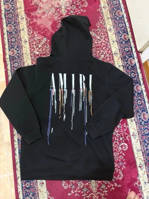 Amiri Hoodie - Säljer en svart hoodie från Amiri med stor logga och färgstänk på ryggen samt mindre tryck på bröstet. Hoodien har huva med dragsko och en klassisk magficka framtill. Perfekt för dig som gillar streetwear och vill sticka ut med cool design.