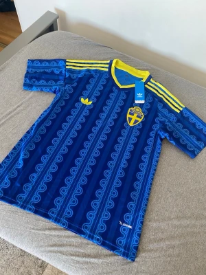 Sverige fotbollströja  - Säljer en blå Sverige tröja från Adidas med gula detaljer och mönster. Tröjan har gula ränder på axlarna, gul krage och Sveriges landslagsemblem på bröstet. Tillverkad i ett lätt och ventilerande material, perfekt för träning eller match.