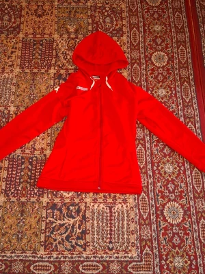 Röd hoodie med dragkedja från Kappa - Snygg röd hoodie från Kappa med dragkedja framtill och vita dragsnören i huvan. Modellen har långa ärmar och två fickor på sidorna. Perfekt för dig som gillar sportig stil och vill ha något bekvämt och stilrent.