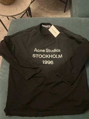 Svart långärmad t-shirt Acne Studios - Svart långärmad t-shirt från Acne Studios med tryck 'Acne Studios STOCKHOLM 1996' på bröstet. Klassisk rund hals och relaxed fit. Perfekt för dig som gillar stilrena och trendiga plagg.