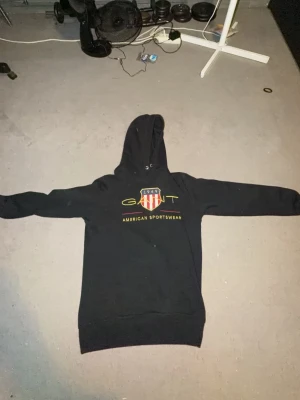 Svart hoodie från GANT med tryck - Säljer en svart hoodie från GANT med broderad logga och amerikanskt sköldmärke på bröstet. Hoodien har dragsko i huvan och ribbade muddar vid ärmslut och nederkant. Står 'AMERICAN SPORTSWEAR' under loggan. Perfekt för en sportig och avslappnad stil.
