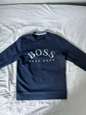 Hugo boss mörkblå  - Snygg mörkblå sweatshirt från Hugo Boss med stort vitt BOSS-tryck på bröstet. Tröjan har rund halsringning och långa ärmar. Perfekt för en clean och stilren look.