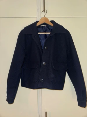 Mörkblå ulljacka med stora fickor - Snygg mörkblå ulljacka med rak passform och stora knappar framtill. Jackan har två rejäla fickor med lock och en klassisk krage. Fodrad insida för extra komfort och värme. Perfekt för kyliga dagar och enkel att matcha med olika outfits.