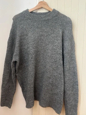 Grå stickad tröja med rund hals - Mysig grå stickad tröja med rund hals och långa ärmar. Tröjan har en enkel, oversized passform och ribbstickade muddar vid hals, ärmslut och nederkant. Perfekt för kalla dagar och enkel att matcha med olika outfits.
