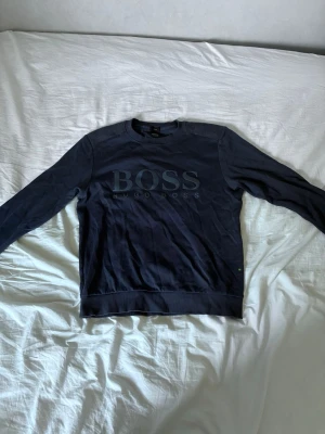 Mörkblå sweatshirt från Hugo Boss - Snygg mörkblå sweatshirt från Hugo Boss med stort tryck på bröstet. Tröjan har rund halsringning, långa ärmar och ribbade muddar vid ärmslut och nederkant. Perfekt för en avslappnad och stilren look.