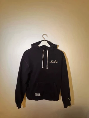 Svart hoodie från New Era - Svart hoodie från New Era med vit broderad logga på bröstet och vita dragsnören i huvan. Klassisk känguruficka framtill och ribbade muddar vid ärmar och nederkant. Perfekt för en avslappnad och stilren look.
