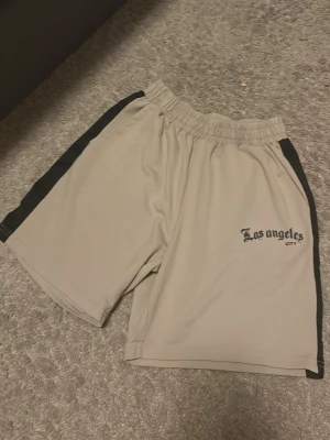 Beige shorts med svarta detaljer - Beiga shorts med svarta paneler längs sidorna och trycket 'Los Angeles City' på vänster ben. De har elastisk midja och är tillverkade i ett syntetmaterial som känns lätt och skönt. Perfekta för träning eller chill dagar på sommaren.