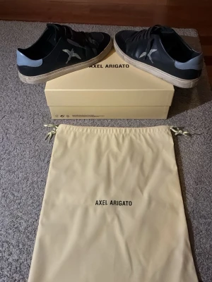 Svarta Axel Arigato sneakers med fågel - Snygga svarta sneakers från Axel Arigato med ljusblå detalj på hälen och broderad fågel på sidan. Skorna har vita sulor och är tillverkade i läder. Perfekta för dig som gillar stilrena och trendiga sneakers. Bra skick fortfarande