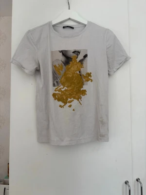 Vit t-shirt med tryck och guldmönster ZARA - Säljer en vit t-shirt med korta ärmar och ett unikt tryck framtill som föreställer ett ansikte och ett abstrakt guldmönster ovanpå. T-shirten har rund hals och är i mjukt bomullsmaterial. Perfekt för dig som vill ha något som sticker ut lite extra. Finns någon fläck