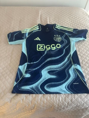 Ajax blå matchtröja Adidas - Säljer en blå Ajax matchtröja från Adidas med ljusblå och neonmönster. Tröjan har korta ärmar, tre ränder på axlarna och Ziggo-logga på bröstet. Materialet är lätt och andas, perfekt för fotboll eller träning.