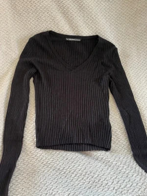 Svart ribbad långärmad tröja - Snygg svart ribbad tröja med lång ärm och djup v-ringning. Tröjan är figurnära och har en croppad passform som passar perfekt till både jeans och kjol. Tillverkad i ett stretchigt material som sitter skönt på kroppen.