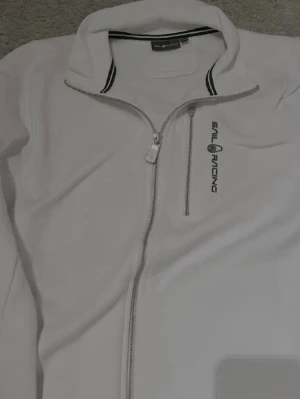 Vit fleecejacka från Sail Racing - Vit fleecejacka med hel dragkedja och hög krage från Sail Racing. Jackan har en bröstficka med dragkedja och svart logotyp på vänster sida. Snygga svarta detaljer på kragen och sportig look.