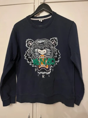 Mörkblå Kenzo sweatshirt med tiger - Säljer en mörkblå sweatshirt från Kenzo med det ikoniska tigermotivet broderat framtill i vitt, grönt och orange. Tröjan har rund halsringning, långa ärmar och ribbade muddar.